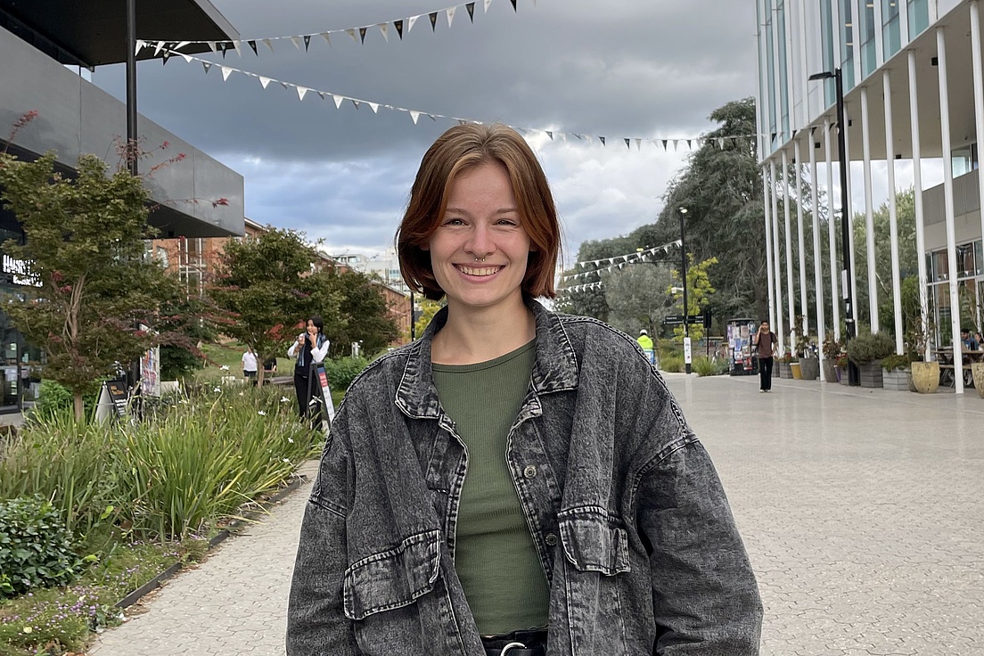 Person mit schulterlangem Haar trägt grüne Bluse und schwarze Jeansjacke steht auf einem Platz mit moderner Architektur und Bäumen im Hintergrund