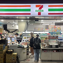 Innenansicht eines 7-Eleven-Ladens mit Kunden an der Kasse und Regalen mit Waren