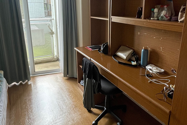 Schlafzimmer mit Einzelbett links, langem Schreibtisch mit Stuhl und Regalen rechts, offener Balkontür mit Vorhängen im Hintergrund