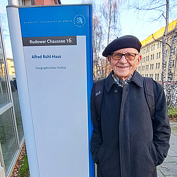 Bernhard Nitz neben einem Schild, dass auf das Alfred Rühl-Haus des Instituts für Geographie hinweist. Er trägt einen dunkelblauen Wollmantel, eine dunkelfarbene Baskenmüthze und einen Rucksack. Er blickt freundlich in die Kamera.