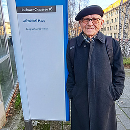 Bernhard Nitz neben einem Schild, dass auf das Alfred Rühl-Haus des Instituts für Geographie hinweist. Er trägt einen dunkelblauen Wollmantel, eine dunkelfarbene Baskenmüthze und einen Rucksack. Er blickt freundlich in die Kamera.