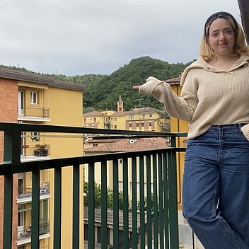 Person lehnt mit einer Hand in der Hosentasche an einem Balkongeländer vor einer Häuserzeile und einem bewaldeten Hügel im Hintergrund