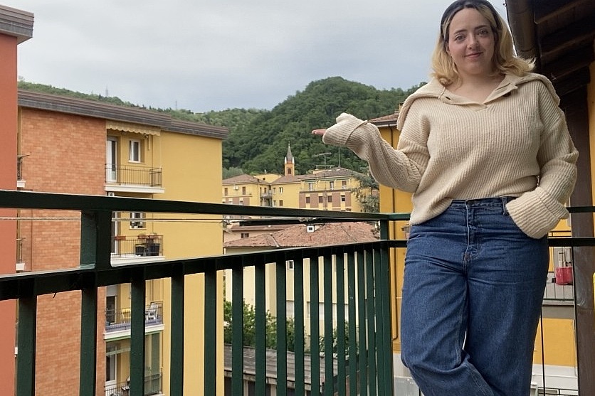 Person lehnt mit einer Hand in der Hosentasche an einem Balkongeländer vor einer Häuserzeile und einem bewaldeten Hügel im Hintergrund