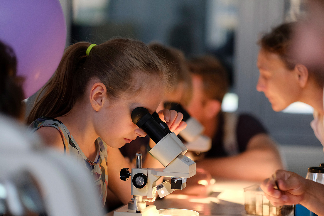 Ein Mädchen schaut durch ein Mikroskop. Neben ihr sind weitere Personen, die das ebenfalls tun. Der Mikroskoptisch ist hell beleuchtet.