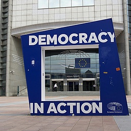 Entrance to a glass building. In front of the entrance is a square blue billboard with the words "Democracy in Action".