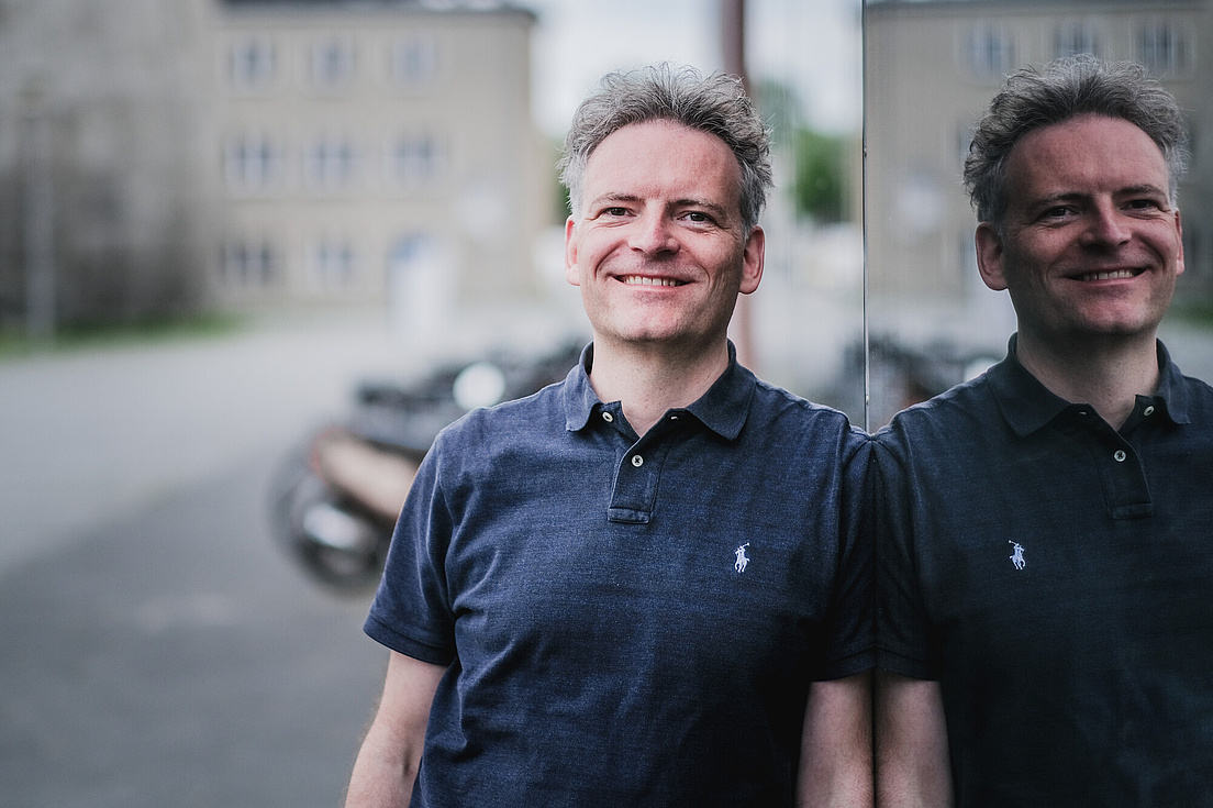 Philipp Adelhelm mit grau meliertem, lockigem Haar steht im Freien und lächelt freundlich in die Kamera. Er trägt ein dunkelblaues Poloshirt mit einem kleinen weißen Pferd-Logo auf der Brust. Der Mann steht vor einer glänzenden, reflektierenden Oberfläche, die sein Spiegelbild zeigt.