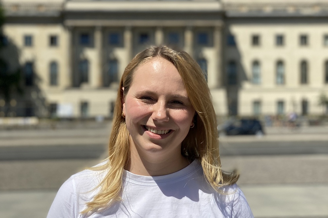 Porträt einer blonden jungen Frau mit strahlendem Lächeln in weißem T-Shirt vor dem Brandenburger Tor mit ukrainischer Flagge unter blauem Himmel.