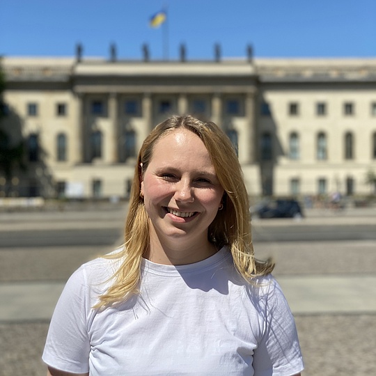 Porträt einer blonden jungen Frau mit strahlendem Lächeln in weißem T-Shirt vor dem Brandenburger Tor mit ukrainischer Flagge unter blauem Himmel.