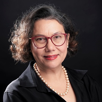 Gökce Yurdakul mit kurzen, gewellten, grau-schwarzen Haaren und roten Brillengläsern blickt freundlich in die Kamera. Sie trägt ein schwarzes Hemd mit aufgestelltem Kragen, eine Perlenkette und hat die Arme vor der Brust verschränkt. Der Hintergrund ist schwarz.