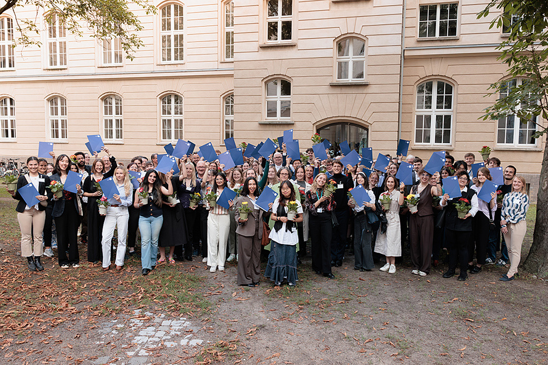 Stipendiat*innen des Deutschlandstipendiums stehen als Gruppe vor einem Gebäude und tragen sommerliche Kleidung. In den Händen halten sie ihre Urkunden hoch.