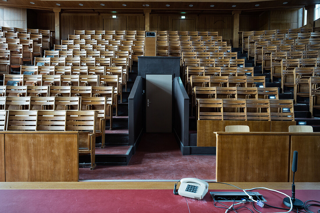 Das Bild zeigt einen leeren Hörsaal oder Vortragssaal mit Holzvertäfelung und Holzsitzen. Die Sitzreihen sind in einem halbkreisförmigen Arrangement angeordnet, wobei die vorderen Reihen tiefer liegen als die hinteren, um eine bessere Sicht für alle Zuhörer zu ermöglichen. In der Mitte des Raumes befindet sich ein Podium mit einer Tür, die vermutlich zu einem Nebenraum führt. Vor dem Podium steht ein Tisch mit einem Telefon, einem Mikrofon und einigen Kabeln darauf. 