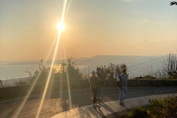 Drei Personen (Mann mit Baby, Frau, Mann) mit Rucksäcken spazieren bei Sonnenuntergang auf einem Aussichtspunkt mit Blick auf Meer, Berge und orange Himmel.