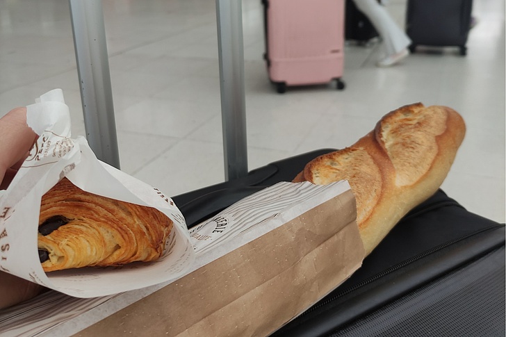 Hände halten Croissant und Baguette in brauner Papiertüte neben pinkem und schwarzem Koffer am Bahnhof.