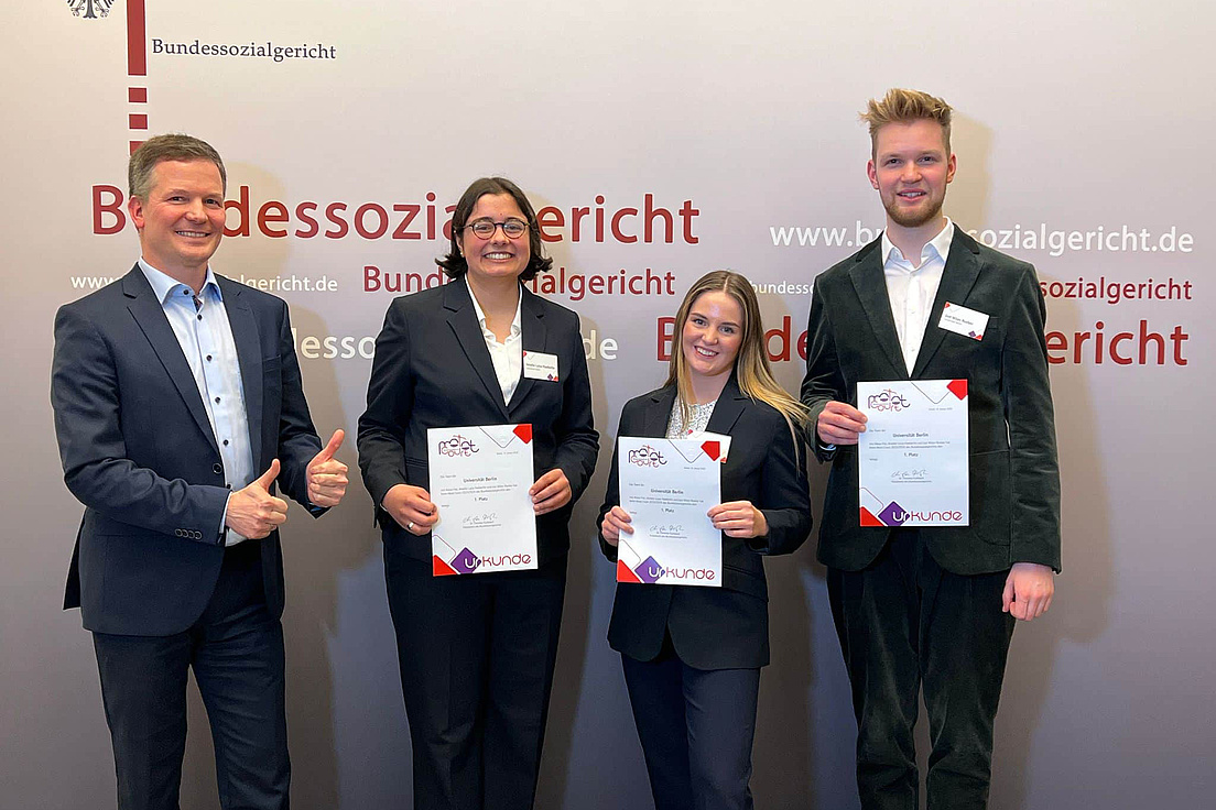Gruppenfoto des Moot-Court Teams der HU: HU-Moot Court Team Amelie Haeberlin, Alexa Frei und Jost Roeber mit Betreuer Dr. Stefan Schifferdecker. Sie tragen alle einen dunkelfarbigen Blazer beziehungsweise einen Anzug, eine dunkelfarbende Hose und ein helles Hemd beziehungsweise Bluse. Sie halten ihre Urkunden und blicken freundlich in die Kamera. Im Hintergrund ist ein helles Plakat mit dem Text Bundessozialgericht und die URL der Webseite des Gerichts zu sehen.