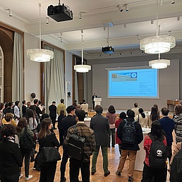 A group of young researchers in a large hall listen to the speech given by the Vice-President of HU.