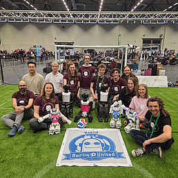 Ein Team posiert mit Robotern auf einem Kunstrasenfeld bei der RoboCupHumanoid Soccer League.