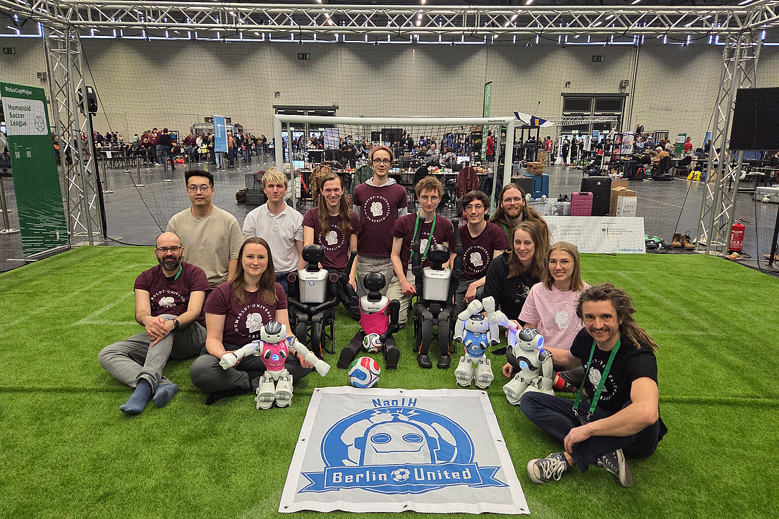 Ein Team posiert mit Robotern auf einem Kunstrasenfeld bei der RoboCupHumanoid Soccer League.