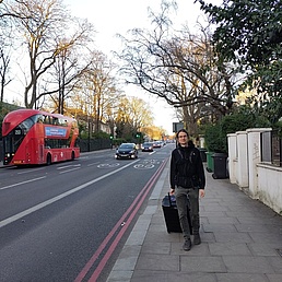  Mann mit Koffer geht bei Sonnenuntergang über Londoner Straße mit rotem Doppeldeckerbus und Bäumen.