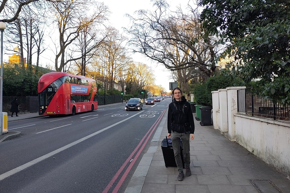  Mann mit Koffer geht bei Sonnenuntergang über Londoner Straße mit rotem Doppeldeckerbus und Bäumen.