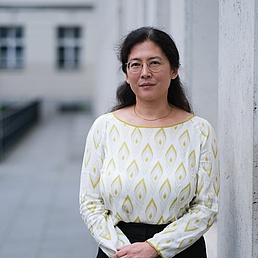 Dr. Yong-Mi Rauch wurde im Freien fotografiert. Links von ihr befindet sich eine graue Wand und im Hintergrund ist ein Gebäude zu sehen. Sie trägt  eine beige Bluse mit einem gelben Muster und einen dunkelfarbenen Rock.