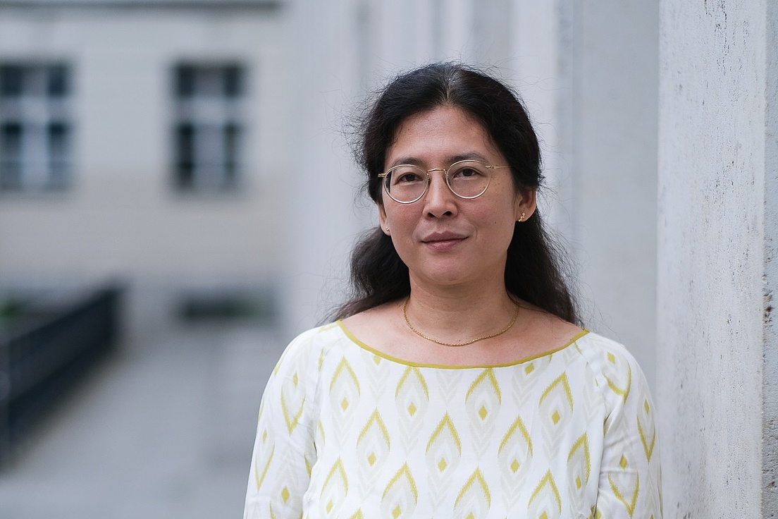 Dr. Yong-Mi Rauch wurde im Freien fotografiert. Links von ihr befindet sich eine graue Wand und im Hintergrund ist ein Gebäude zu sehen. Sie trägt  eine beige Bluse mit einem gelben Muster und einen dunkelfarbenen Rock.