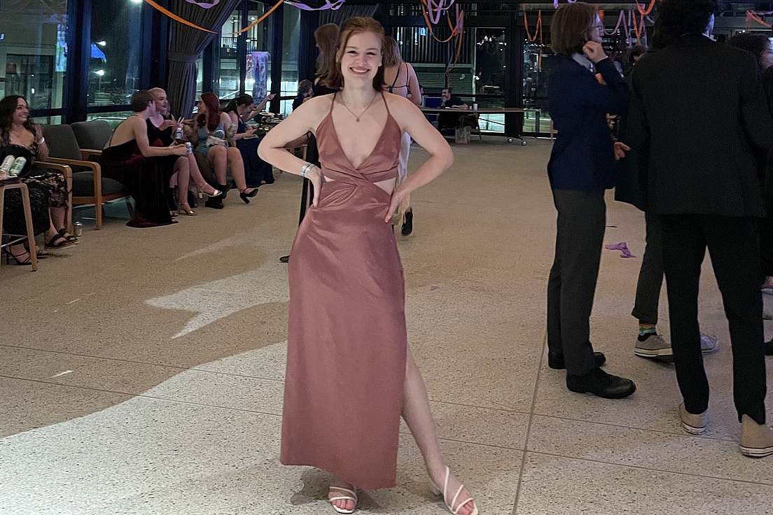 Frau in langem, ärmellosem Abendkleid mit Spaghettiträgern und Schlitz steht mit Händen in der Taille in einem Innenraum mit mehreren Personen im Hintergrund.