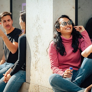 Gruppe von Studierenden unterhält sich entspannt im Freien, eine Frau in einem pinken Pullover lächelt.