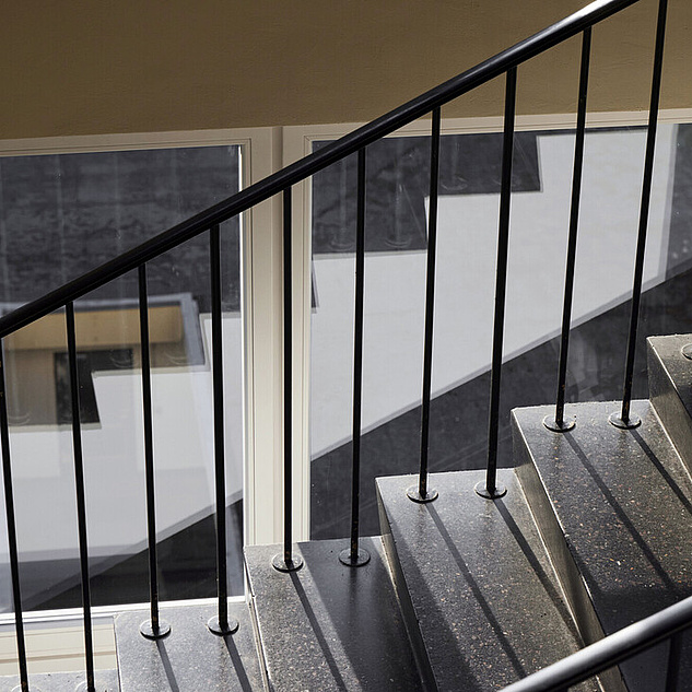 Forschung-Lehre-Berufungen-Wissenschaftliche-Karriere-Campus-Mitte-20220806-Julia-Baier Das Bild zeigt eine Treppe mit schwarzem Geländer und Stufen aus glänzendem Material, vermutlich Granit oder Marmor. Die Treppe führt nach unten zu einem beleuchteten Raum mit großen Fenstern. Durch die Fenster ist ein Teil eines Gebäudes mit einer weißen Fassade und dunklen Fenstern zu sehen. Das Licht fällt durch die Fenster und beleuchtet den Raum und die Treppe.