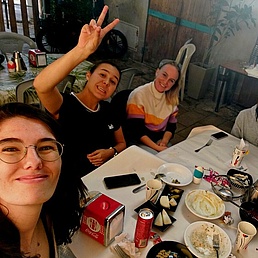 Gruppe von Menschen sitzt an einem Tisch mit Tellern, Getränkedosen und Bechern in einem Innenraum mit Holzwänden.