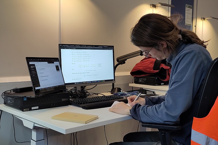 Herr mit Brille und Pferdeschwanz in blauem Hoodie sitzt konzentriert an einem hellen Schreibtisch mit zwei Monitoren (Code), Laptop, Notizblock und Stift in einem Büro mit Lampe und oranger Sicherheitsweste
