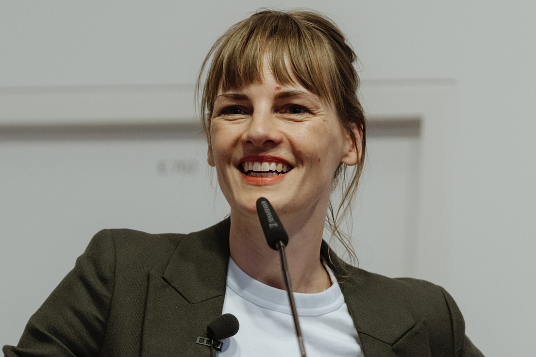 Romy Jasper steht hinter einem Rednerpult. Vor ihr ist ein Mikrofon und sie blickt freundlich in die Kamera.