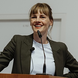 Romy Jasper steht hinter einem Rednerpult. Vor ihr ist ein Mikrofon und sie blickt freundlich in die Kamera.