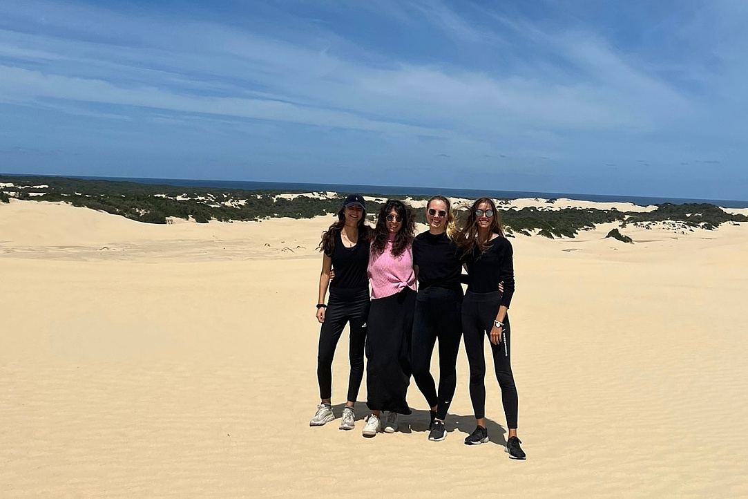 Vier Frauen in legerer Sommerkleidung mit Cap und Sneakers posieren lachend auf Sanddünen mit Ozean und blauem Himmel im Hintergrund.