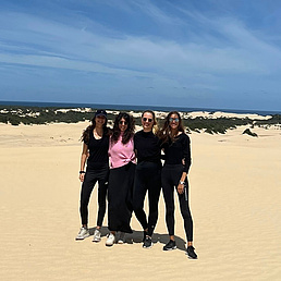 Vier Frauen in legerer Sommerkleidung mit Cap und Sneakers posieren lachend auf Sanddünen mit Ozean und blauem Himmel im Hintergrund.