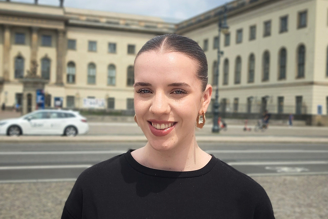 Junge Frau mit blondem Pferdeschwanz, großen Gold-Ohrringen und schwarzem Pullover lächelt vor historischen Gebäuden mit Säulen und einer weißen Limousine auf der Straße im Hintergrund.