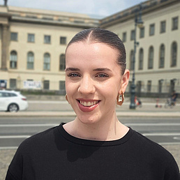 Young woman with blonde ponytail, large gold hoop earrings, and black sweater smiles in front of historic buildings with columns and a white limousine on the street in the background.