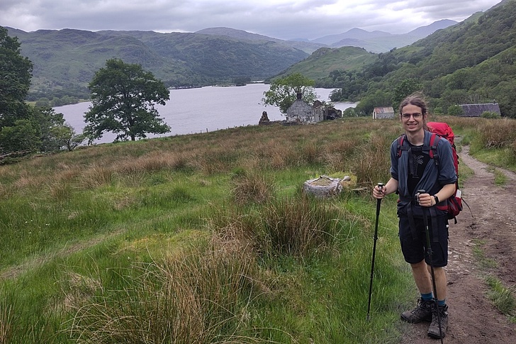Mann mit Trekkingrucksack und Stock wandert bei regnerischem Wetter durch grüne Hügel mit See und Bergen im Hintergrund