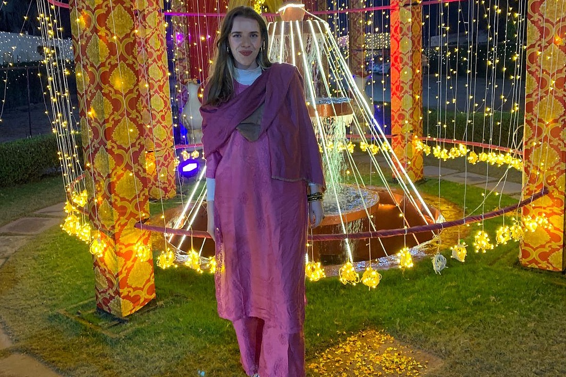 Frau mit langen Haaren in pinkvioletter traditioneller indischer Salwar-Kameez steht abends vor einem beleuchteten, mit Lichterketten und Girlanden geschmückten Springbrunnenpavillon auf Rasen.