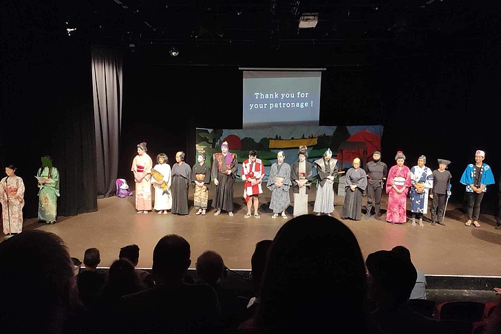 Personen in traditionellen japanischen Kostümen stehen auf einer Bühne vor einem Publikum, im Hintergrund ein Bildschirm mit der Aufschrift 'Thank you for your patronage'