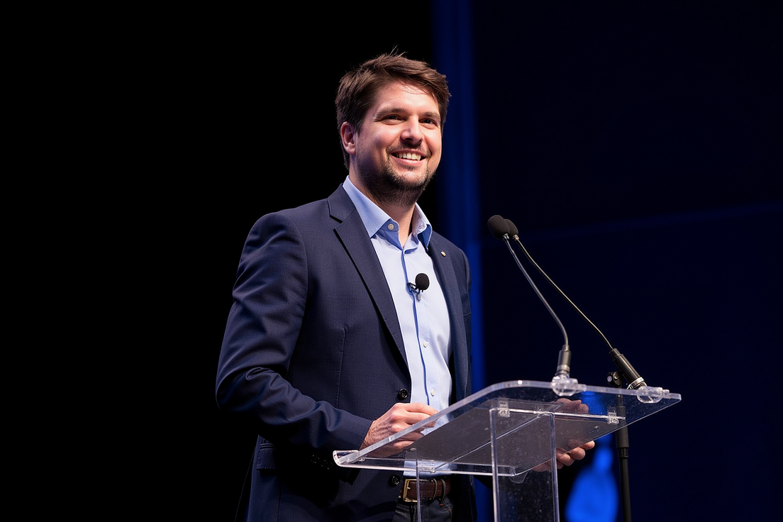 Open Science Ambassador Matthias König steht vor einem Rednerpult mit Mikrofon und lächelt ins Publikum. Er trägt ein hellblaues Hemd und ein dunkelblaues Sakko.