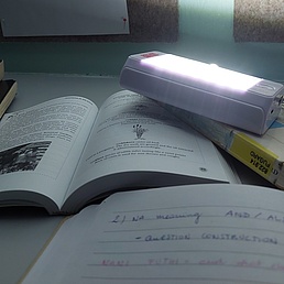 Aufgeschlagene Bücher und ein Notizbuch mit handschriftlichen Notizen auf einem Tisch, darüber eine leuchtende Schreibtischlampe.