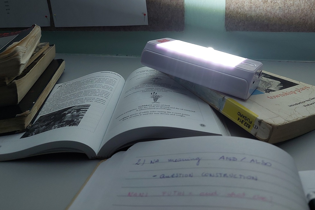 Aufgeschlagene Bücher und ein Notizbuch mit handschriftlichen Notizen auf einem Tisch, darüber eine leuchtende Schreibtischlampe.