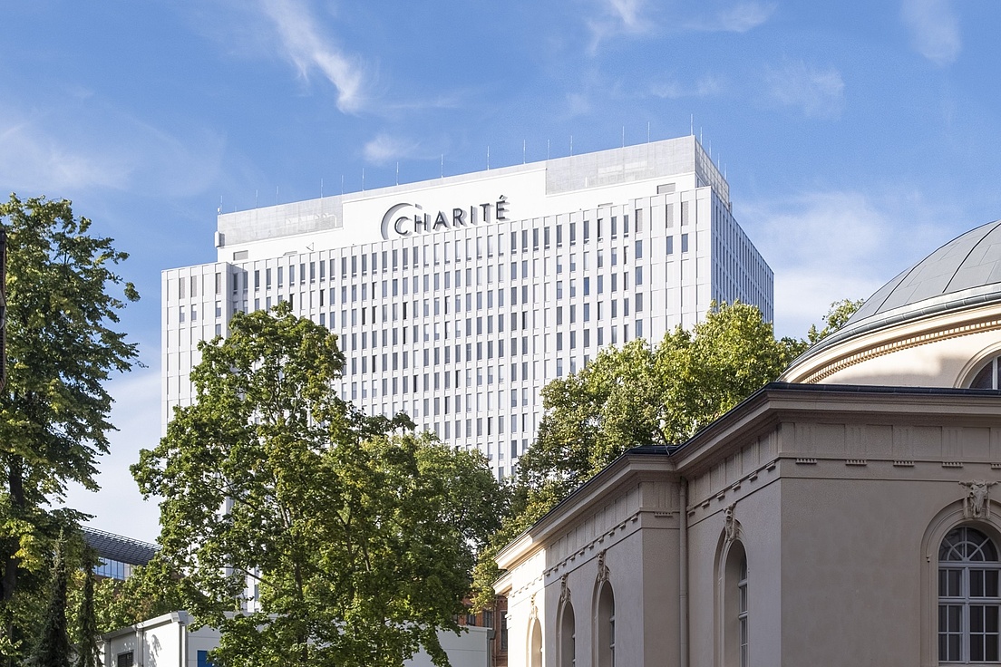Modern Charité building in Berlin with a white facade and grid-like windows. In the foreground, there is a historic structure with a dome. The sky is partly cloudy.
