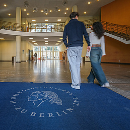 Zwei Personen laufen über einen blauen Teppich mit HU-Siegel, der am Eingang der Juristischen Fakultät liegt, drüber.