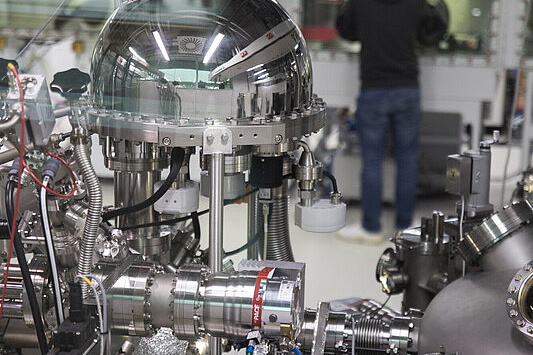 Open-Humboldt-Forschung, Campus Adlershof Das Bild zeigt ein hochkomplexes wissenschaftliches Gerät, vermutlich ein Teil eines Vakuum- oder Hochvakuum-Systems, das in einem Labor eingesetzt wird. Im Vordergrund ist ein glänzender, halbkugelförmiger Behälter zu sehen, der aus Metall gefertigt ist und an mehreren Stellen mit Schläuchen, Kabeln und anderen technischen Komponenten verbunden ist.