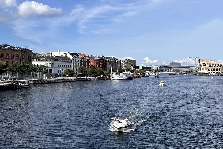 Fluss mit Booten und Häuserzeile am Ufer unter blauem Himmel mit Wolken