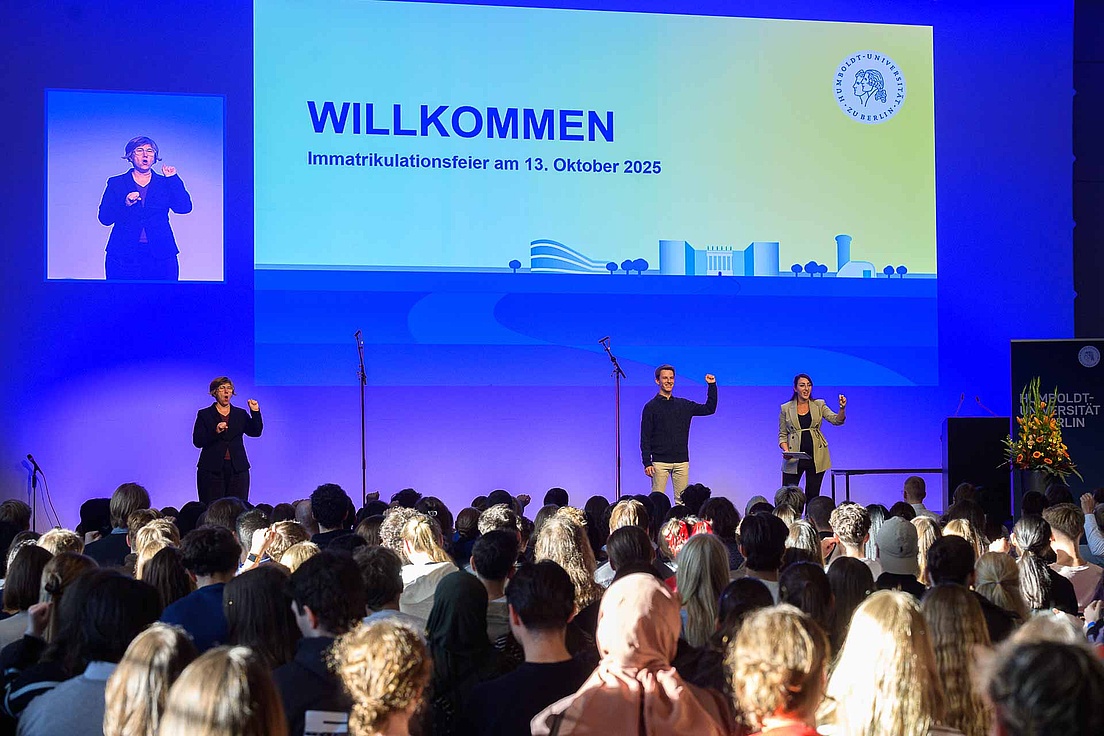 Immatrikulationsfeier 2025 im Humboldt Labor Publikum, Moderaror*innen und Gebärdendolmetscherin bei der Immafeier. Auf einem Bildschirm steht ua. Willkommen