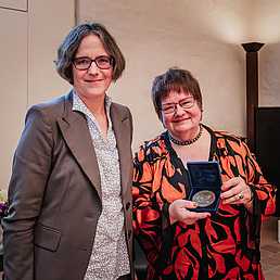 Dr.in Ursula Fuhrich-Grubert hält die Humboldt-Medaille in die Kamera und lächelt. Sie trögt eine orange-schwarz gemusterte Jacke und ein schwarzes Shirt. Julia von Blumentahl steht neben ihr und trägt einen taupe-farbenen Blazer und eine weiß-schwarz gemusterte Bluse.