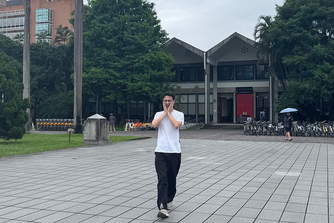 Person in weißem T-Shirt und dunkler Hose steht auf gepflastertem Platz vor einem zweistöckigen Gebäude mit spitzem Dach und roten Türen.