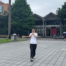 Person in weißem T-Shirt und dunkler Hose steht auf gepflastertem Platz vor einem zweistöckigen Gebäude mit spitzem Dach und roten Türen.